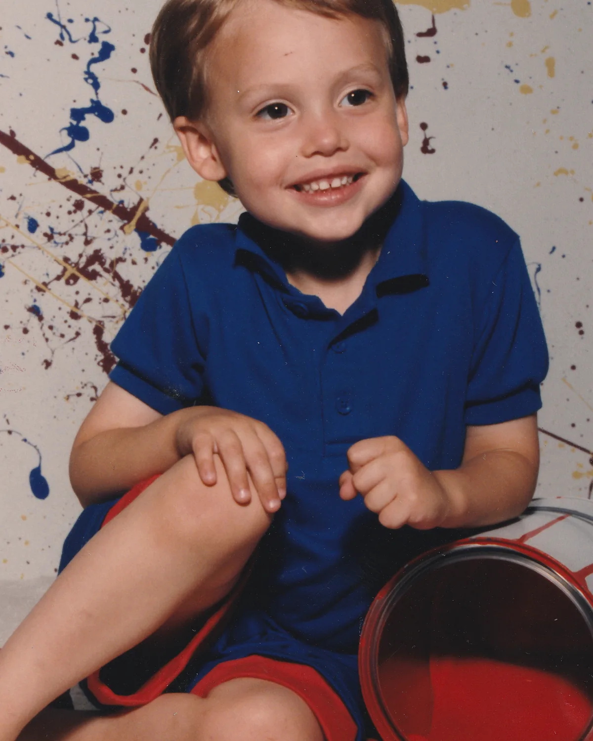 Scott Hancock as a small boy, sitting on a paint-spattered floor in a blue shirt beside an open can of red paint.