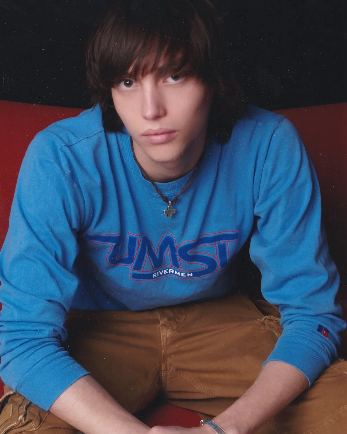 Scott Hancock as a young man, seated on a red couch in a University of Missouri–St. Louis Rivermen sweatshirt, wearing a cross on a chain.